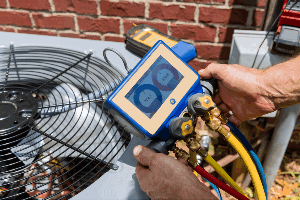 HVAC technician checks refrigerant pressure on an outdoor air conditioning condenser using a digital manifold gauge connected to color-coded hoses.